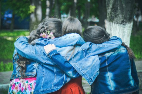 three girl friends wearing denim jackets facing away with their arms around each other as they look towards trees