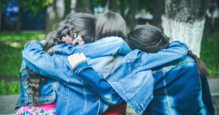 three girl friends wearing denim jackets facing away with their arms around each other as they look towards trees